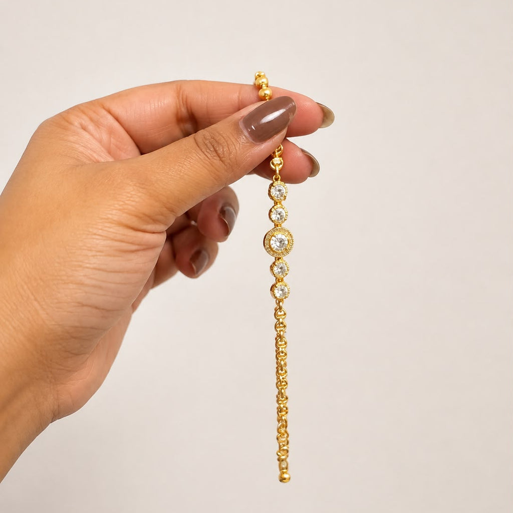 A gold-toned beaded chain earring with a central round bezel-set clear stone, held by a hand against a plain light beige background.