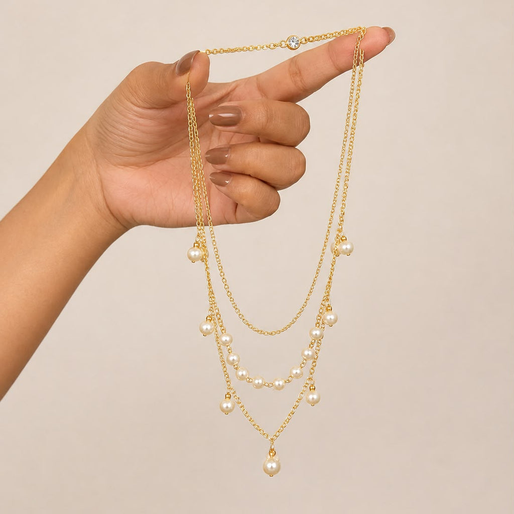A gold-toned multi-layer necklace with small pearl-like beads and a larger pearl pendant, held up by a hand against a plain beige background.