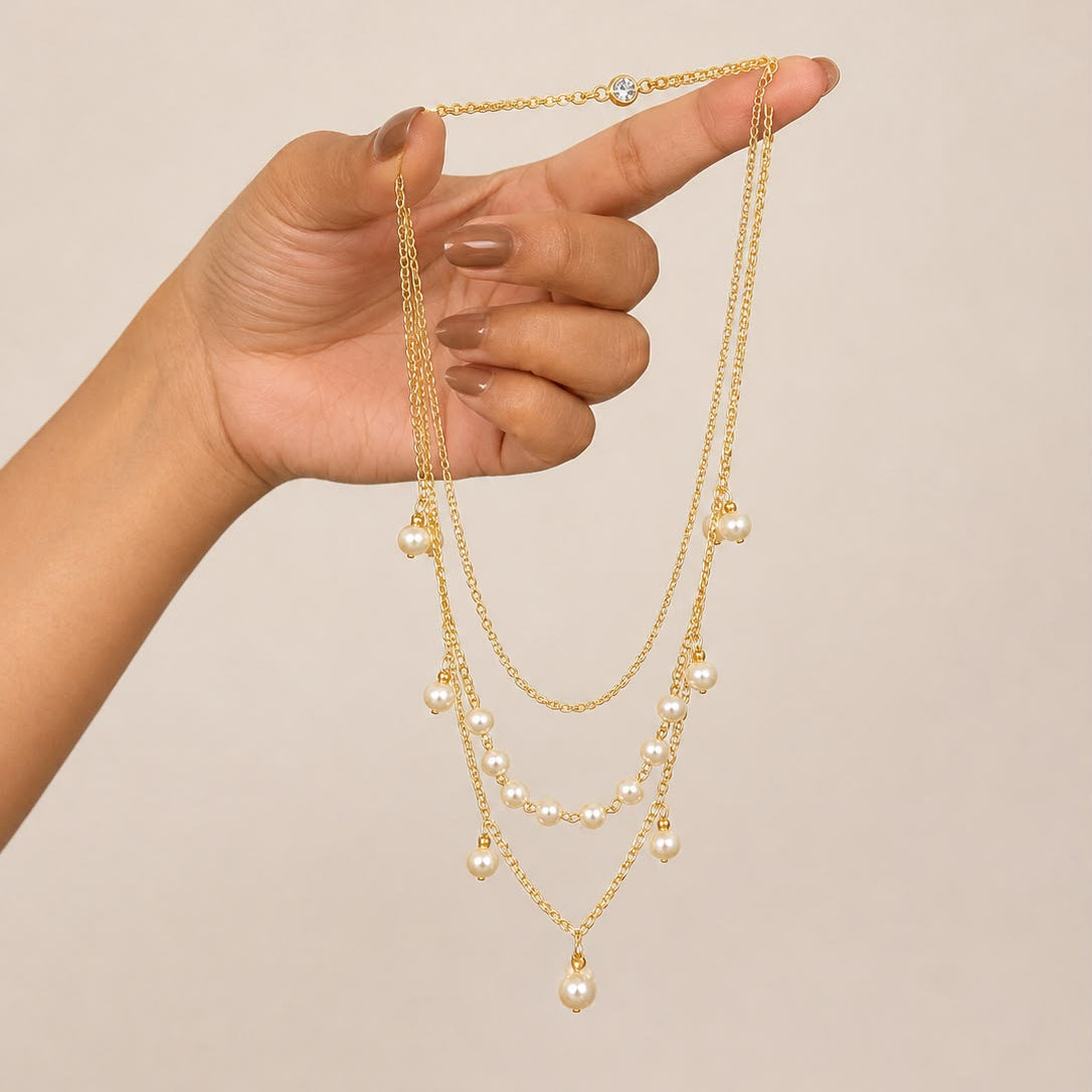 A gold-toned multi-layer necklace with small pearl-like beads and a larger pearl pendant, held up by a hand against a plain beige background.